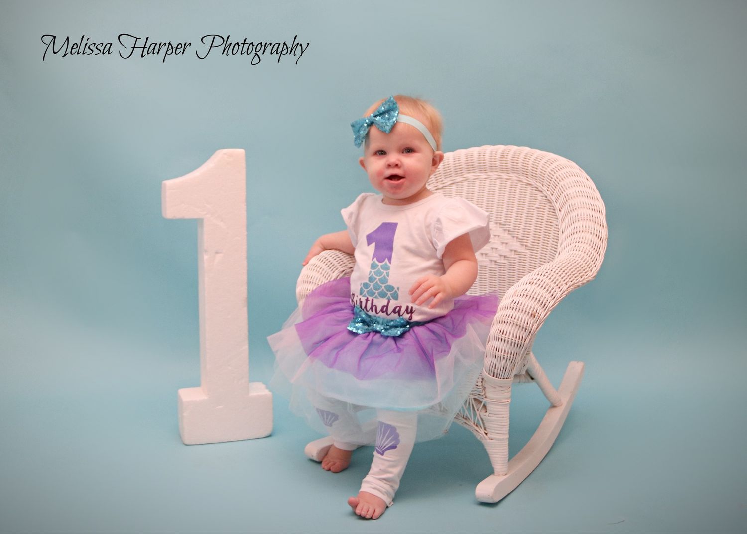 First birthday photo setup with large white number one and wicker rocking chair against blue backdrop.