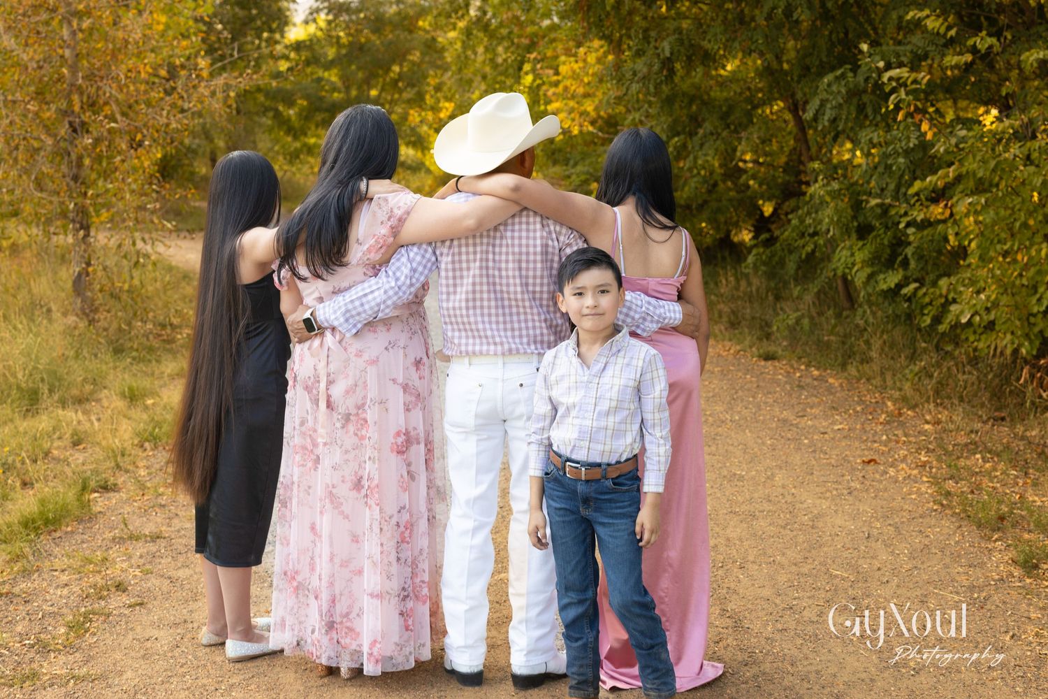 Group poses on a dirt trail surrounded by autumn foliage wearing pink dresses, western attire, and dark outfits.