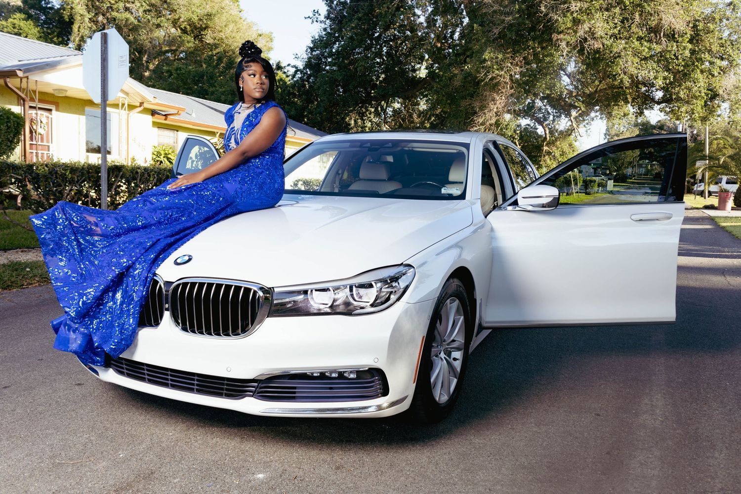 Young woman in royal blue gown posing elegantly beside a white BMW luxury sedan outdoors.