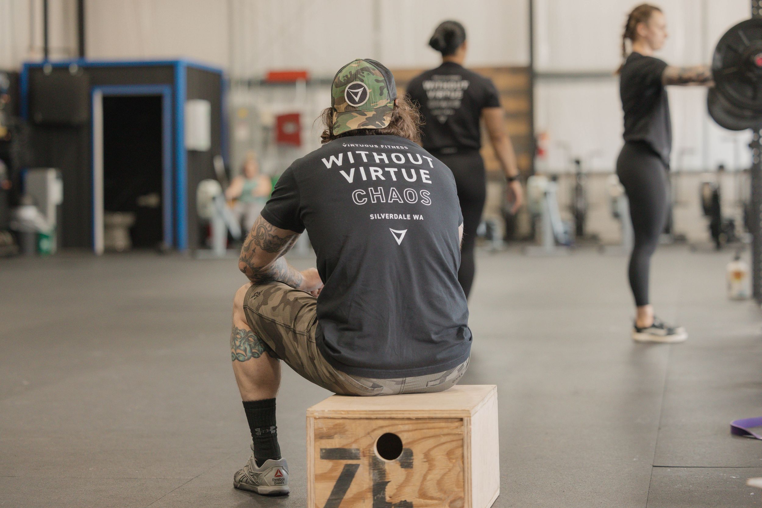 Person in black shirt and camo hat sitting on wooden box in gym setting.