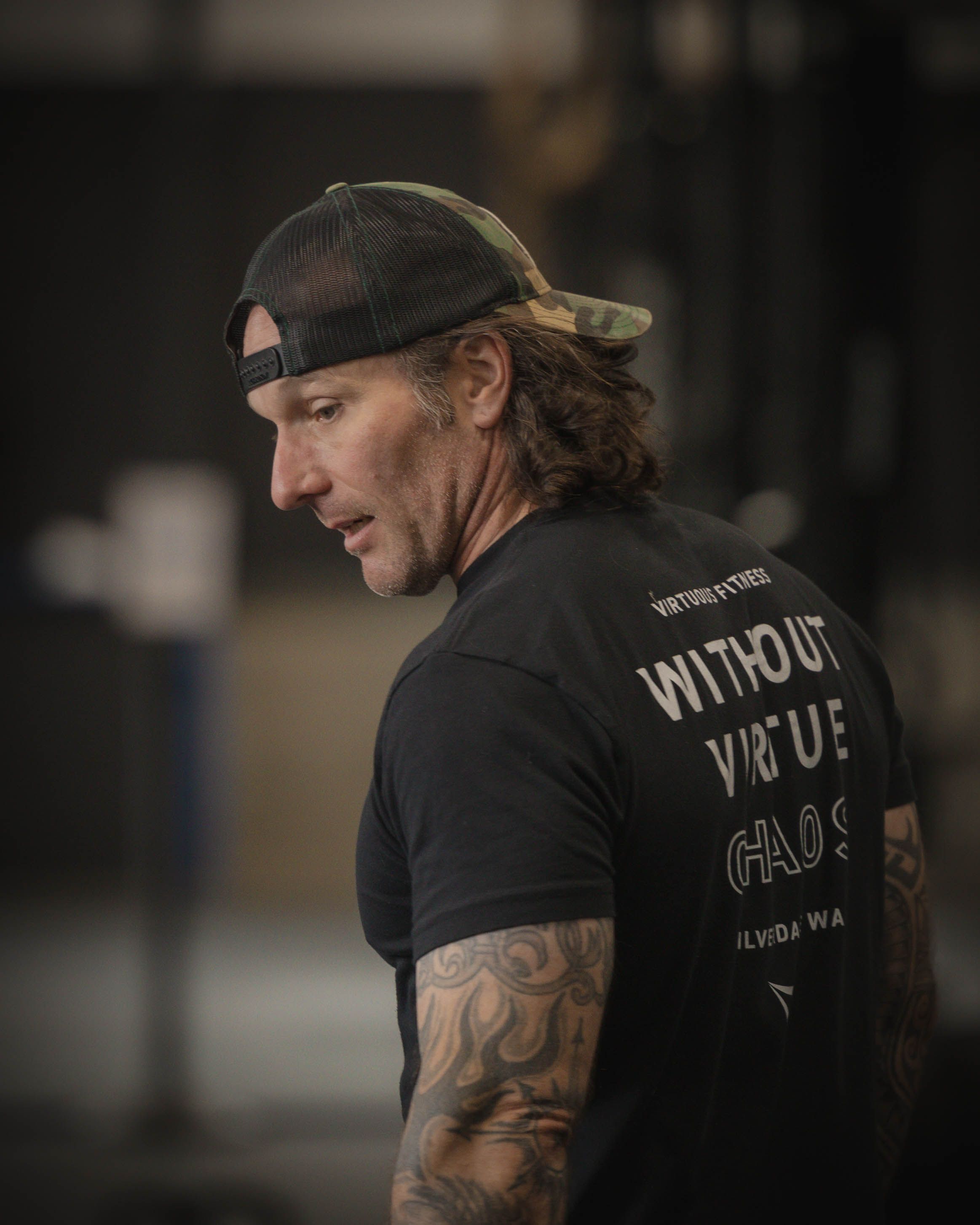 Person wearing black t-shirt and camo hat with Without Virtue text in indoor gym setting.