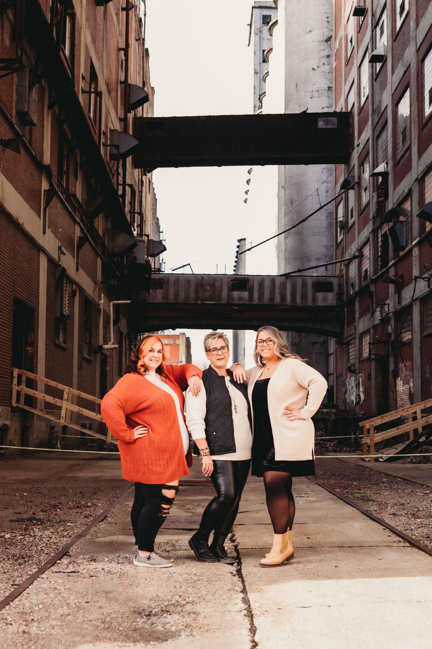 Three friends pose in winter sweaters in an urban alleyway with industrial overhead bridges.