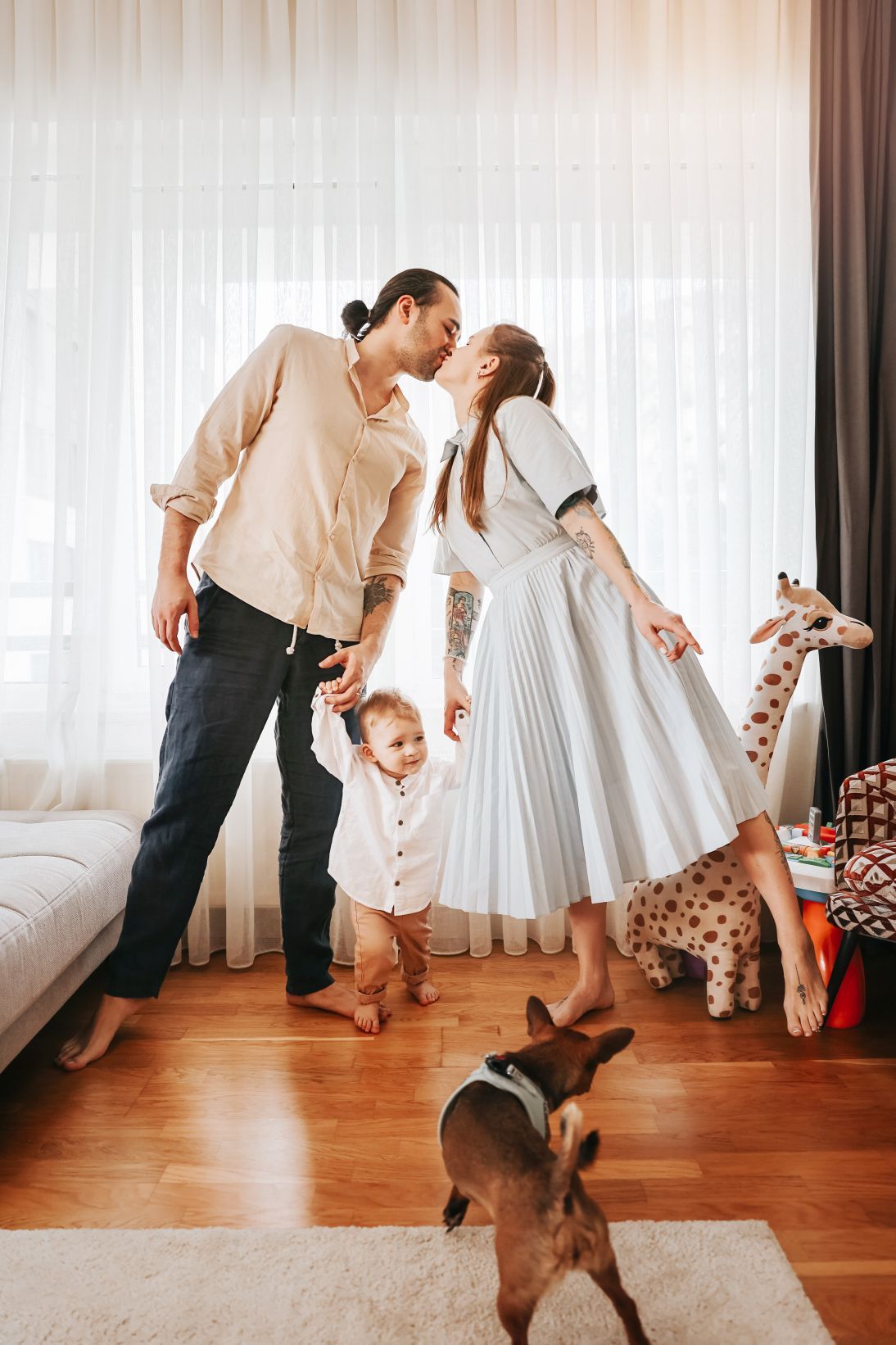 A sweet family moment at home with toys and a white pleated dress against sheer curtains.