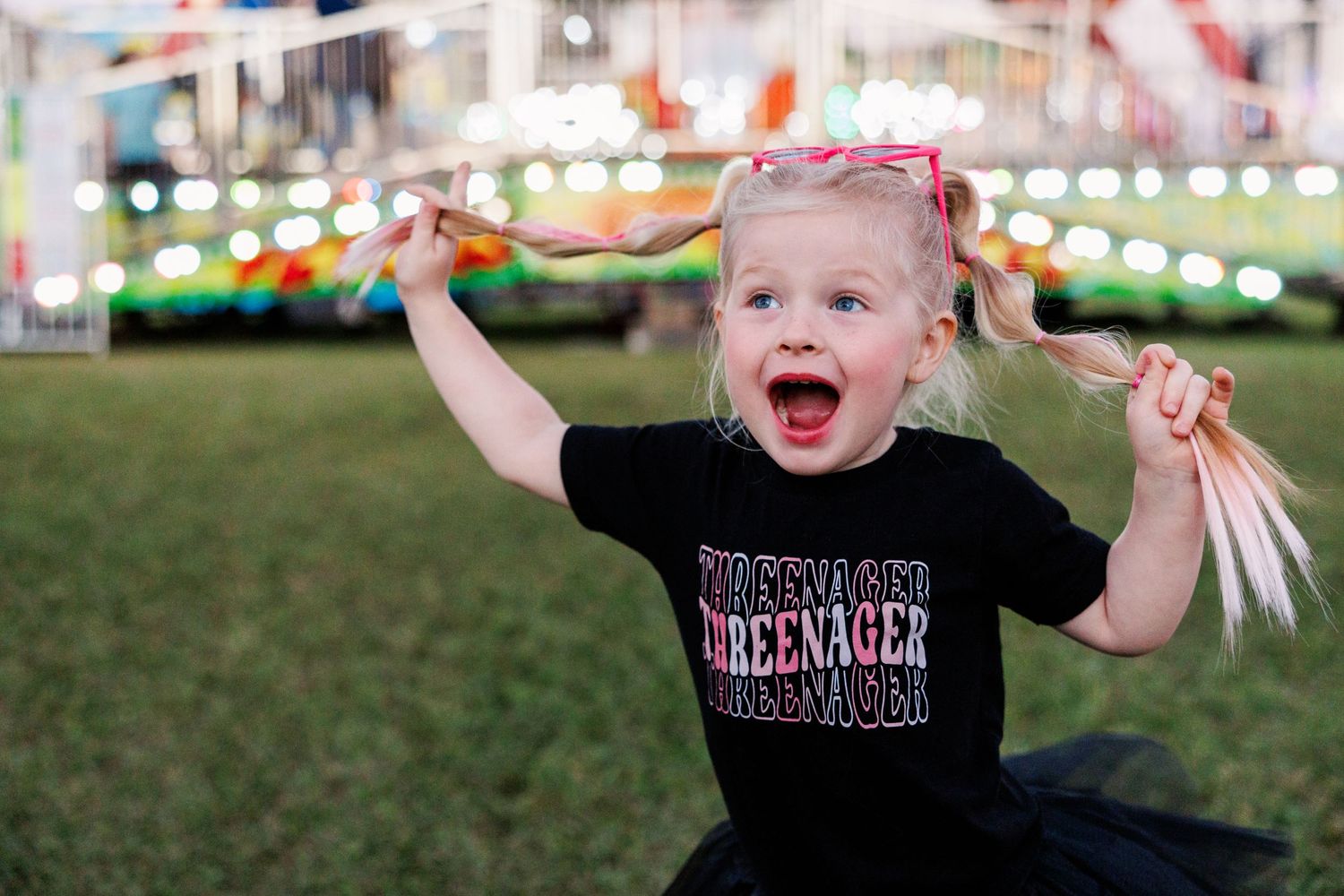 Small child in black Zeppelin shirt at carnival smiles with joy while holding their pigtails up in the air.