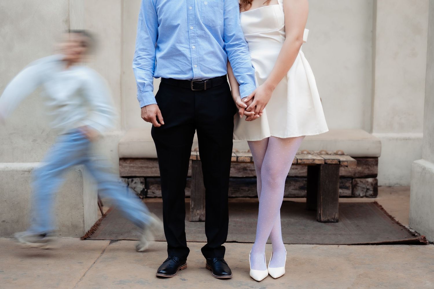 A couple stands together while a blurred figure runs past, with the woman in a pink dress and man in blue shirt and dark pants.