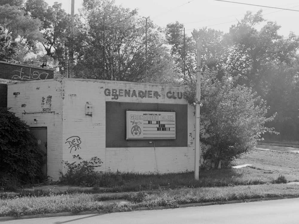 Black and white photograph of an abandoned Grenadier Club building exterior with overgrown vegetation.
