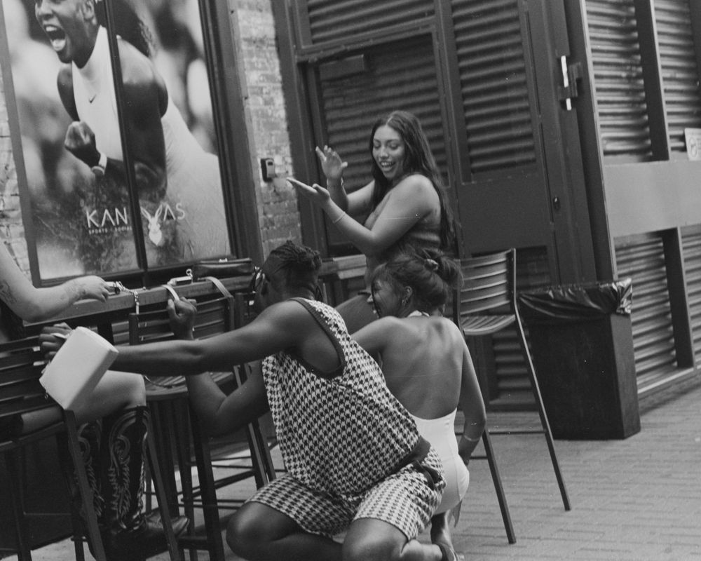 Black and white photo of people seated at outdoor cafe tables having conversation and gesturing in an urban setting.