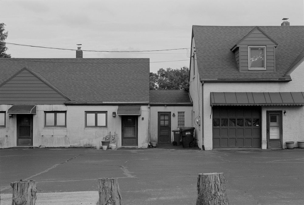 Black and white photograph of two connected buildings with garage doors and tree stumps in foreground.