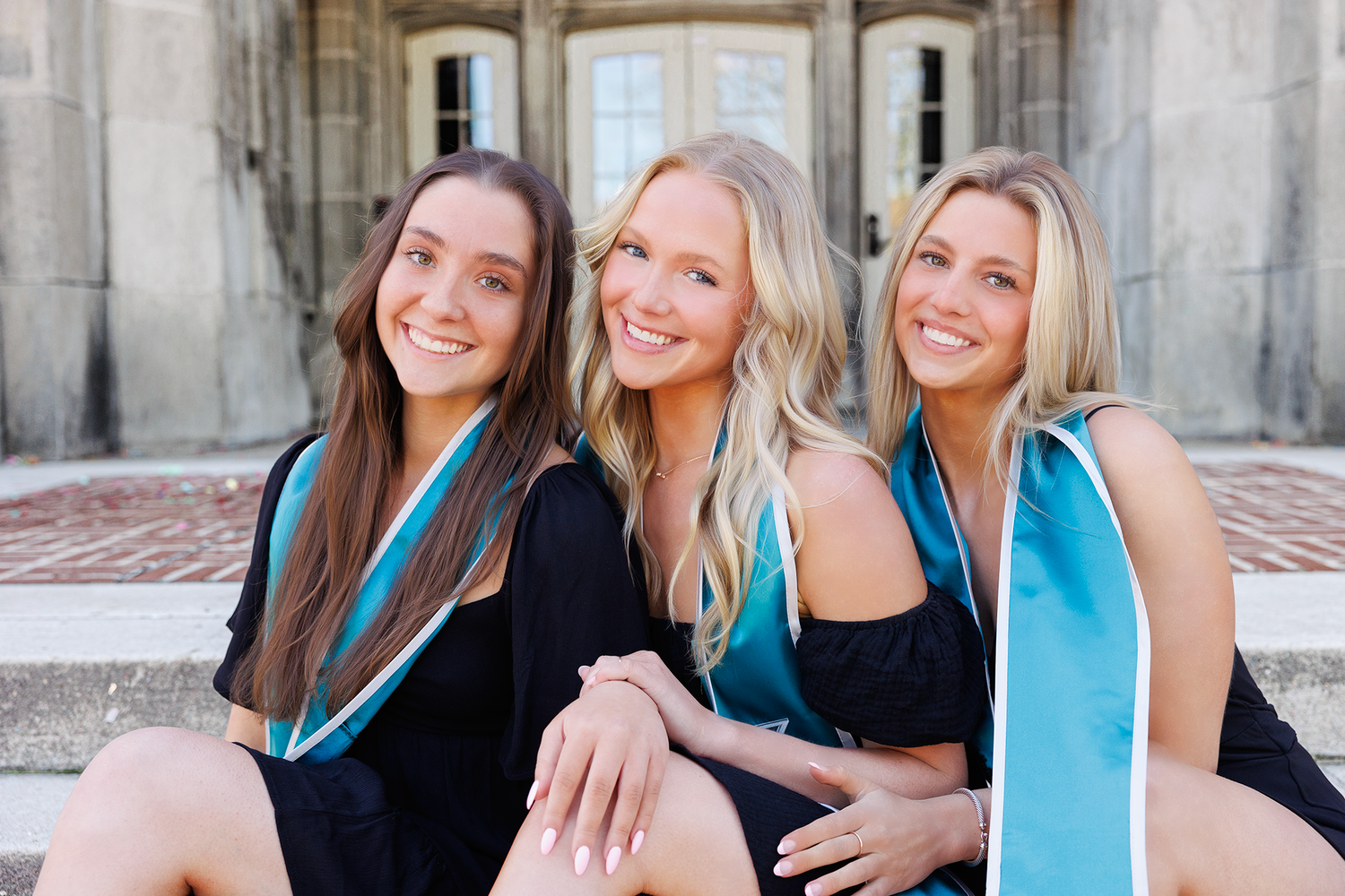 Three graduates wearing black dresses and turquoise stoles sit together smiling during commencement celebration.