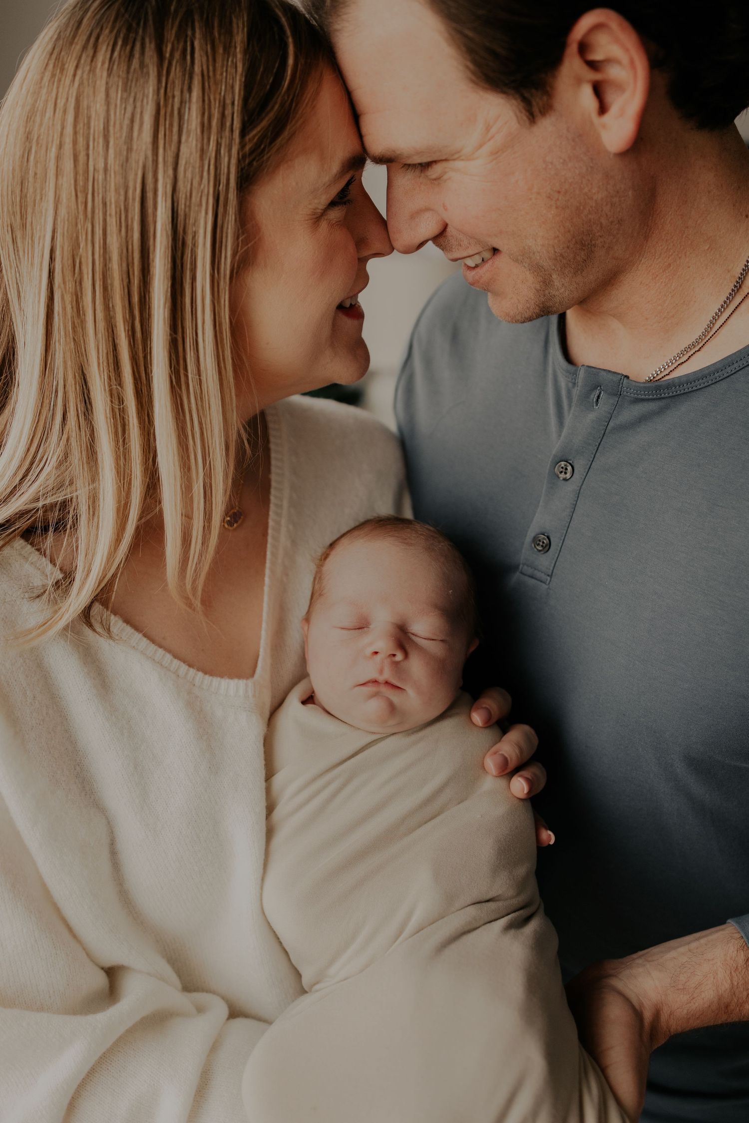 Intimate family portrait showing parents sharing tender moment while holding their sleeping newborn baby in soft lighting.