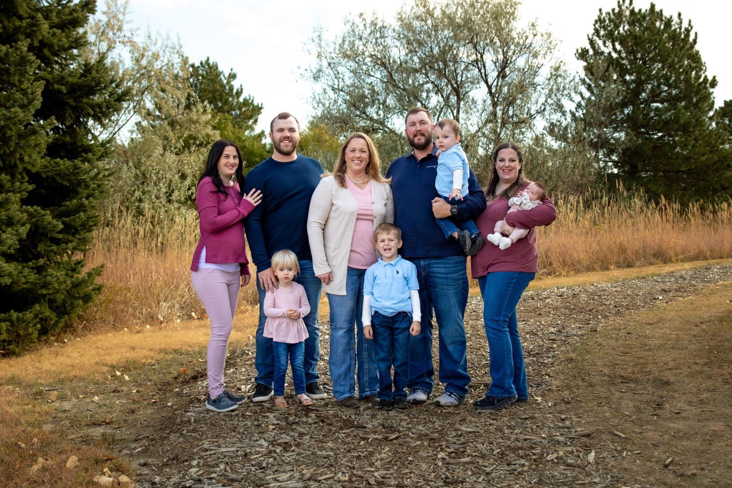 Extended family poses together on a dirt path surrounded by pine trees and golden grass during sunset.