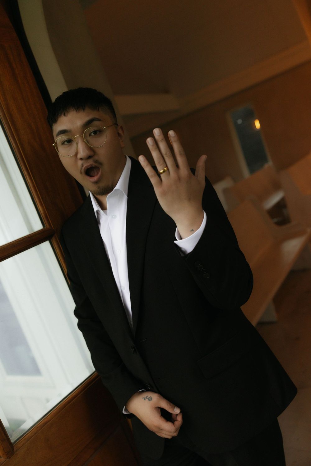 Groom in black suit showing off wedding ring with surprised expression near a window indoors.