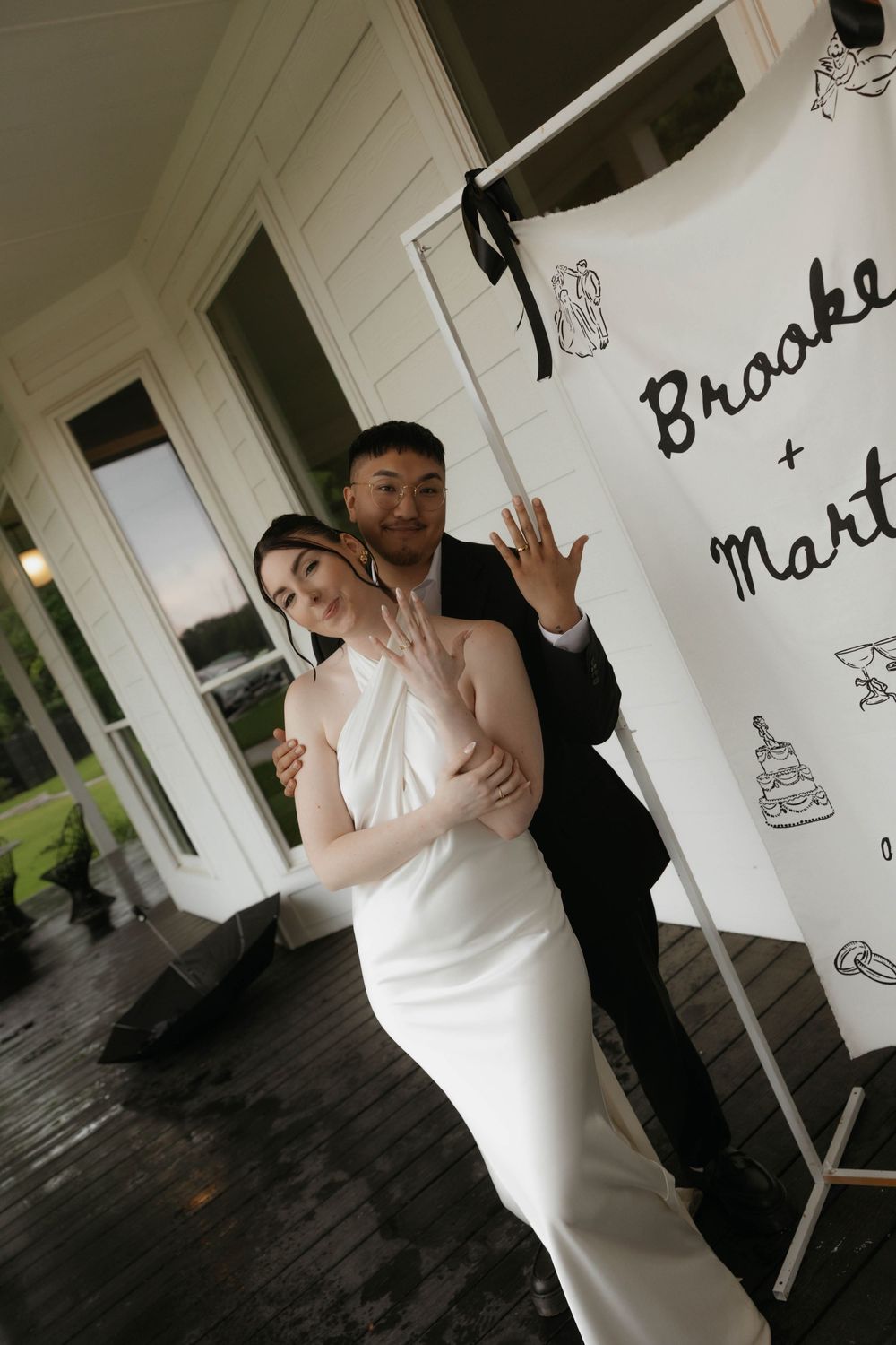 Newlyweds pose showing wedding rings beside personalized Brooke and Martin wedding banner on porch.