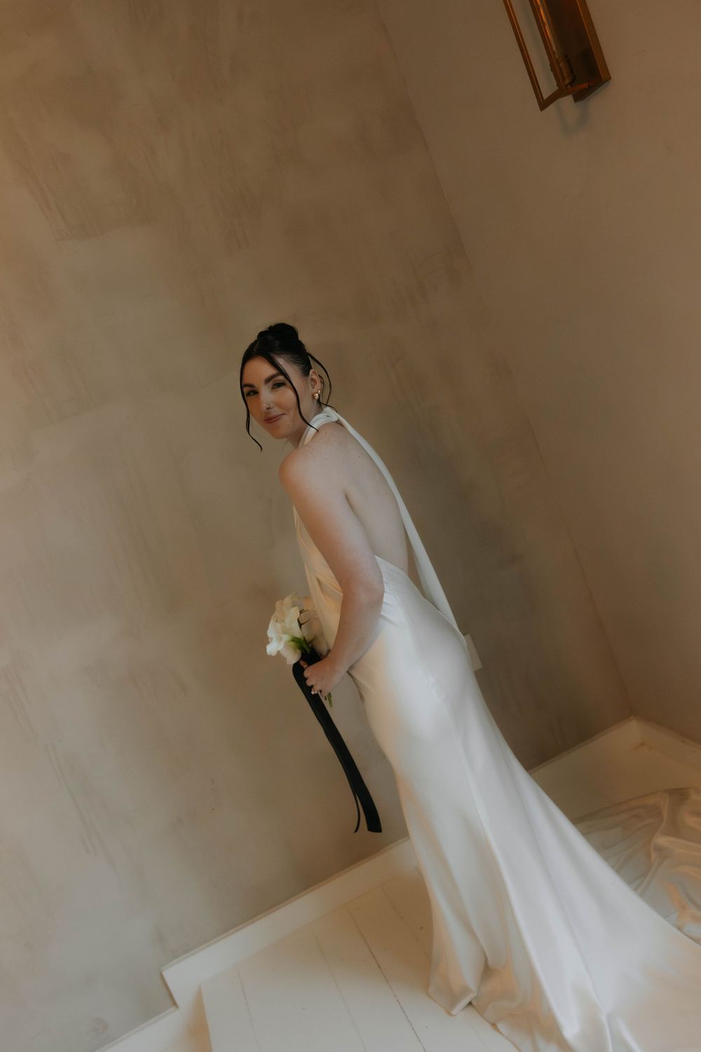 Bride in a white halter neck satin gown holding a bouquet while standing on stairs.