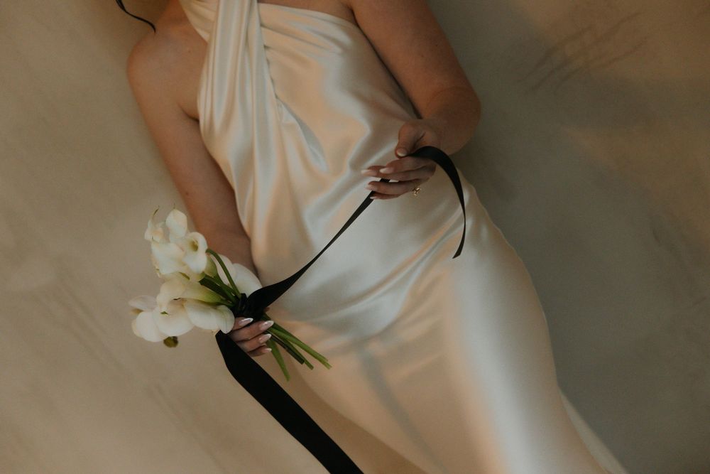 Close-up of a bride in a white satin halter gown holding white calla lilies with black ribbon.