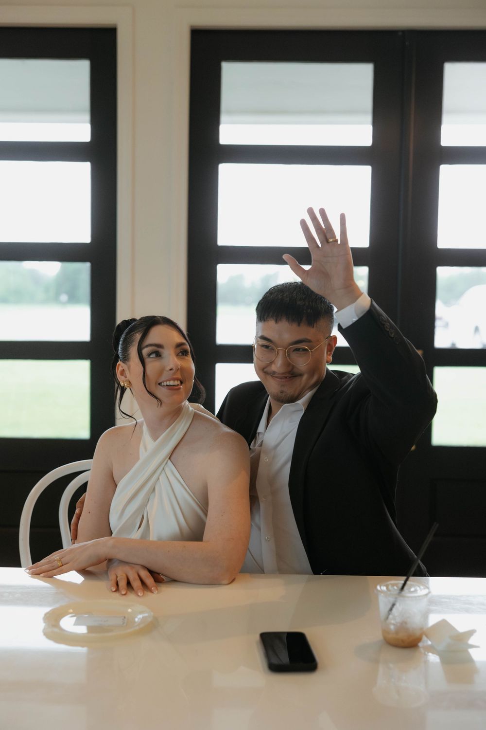 Bride and groom sitting at a white table, groom raising hand showing wedding ring, smiling joyfully.