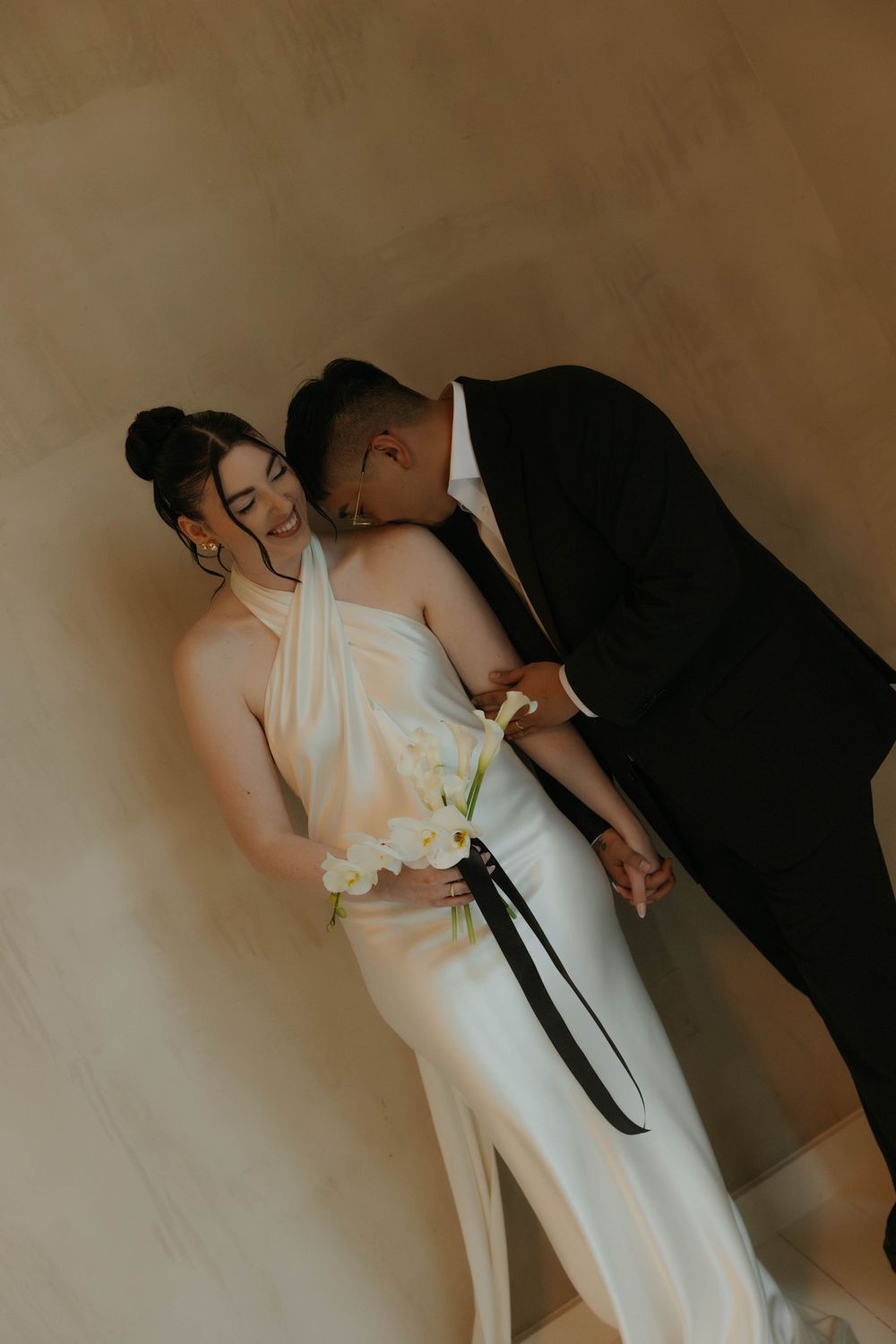 Bride in white halter gown holding calla lilies while groom in black suit kisses her cheek against beige wall.