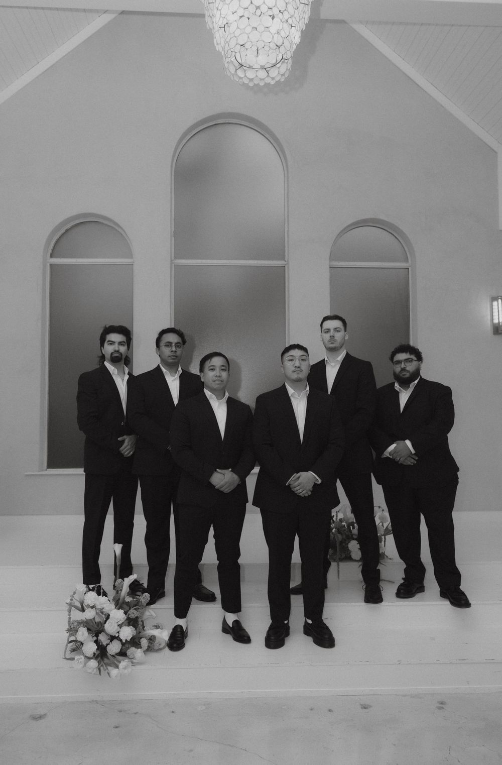 Black and white portrait of groom with five groomsmen in black suits standing in elegant chapel venue.