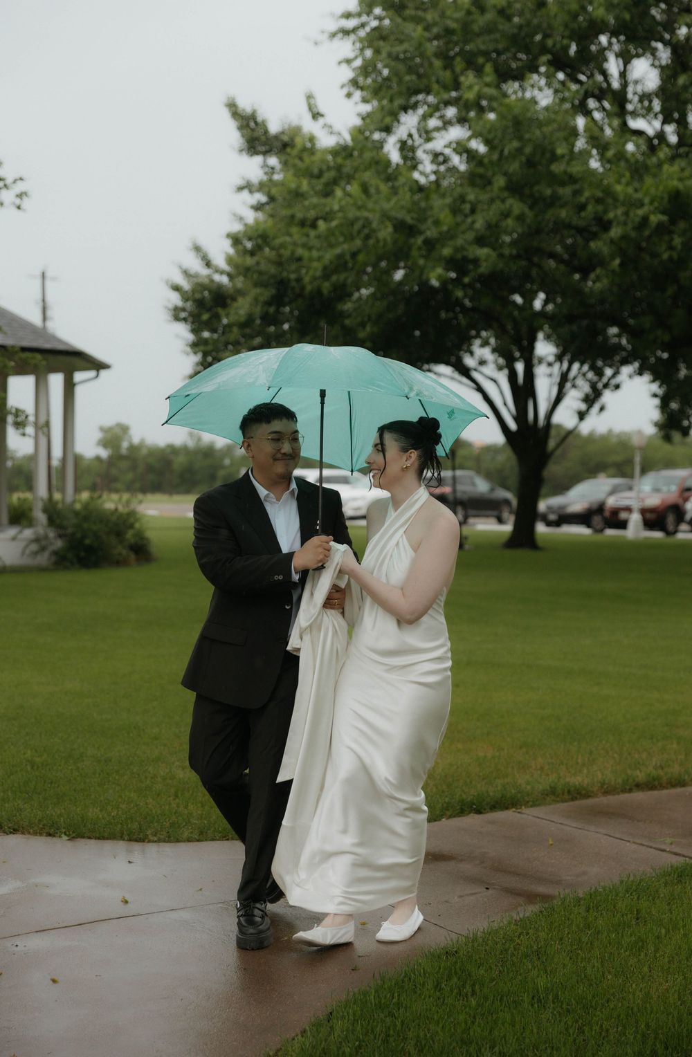 Bride and groom sharing a teal umbrella in the rain on their wedding day outdoors.