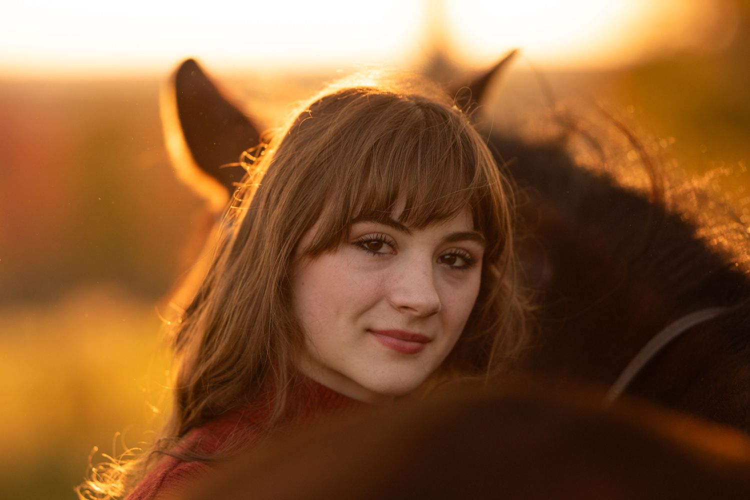 Young woman with bangs smiling warmly beside a dark horse at golden sunset. Photo by Xenophon Photography in Maine.