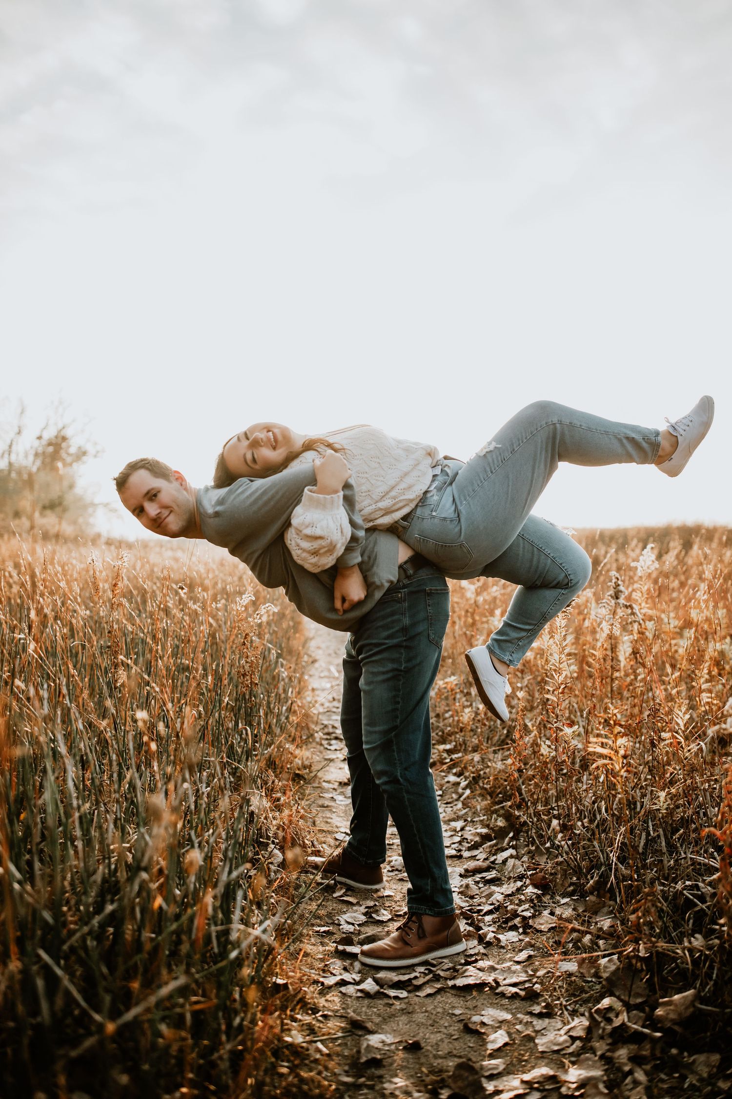 A candid moment shows a playful couple enjoying a fall day on a grassy path at sunset.