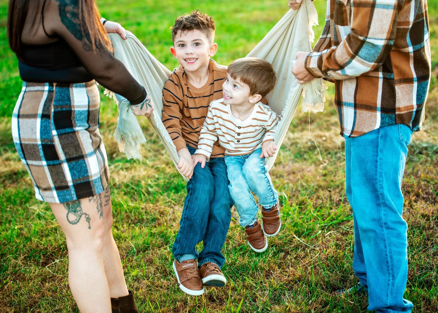 Two children sitting on a blanket swing while adults stand nearby in a grassy outdoor setting.
