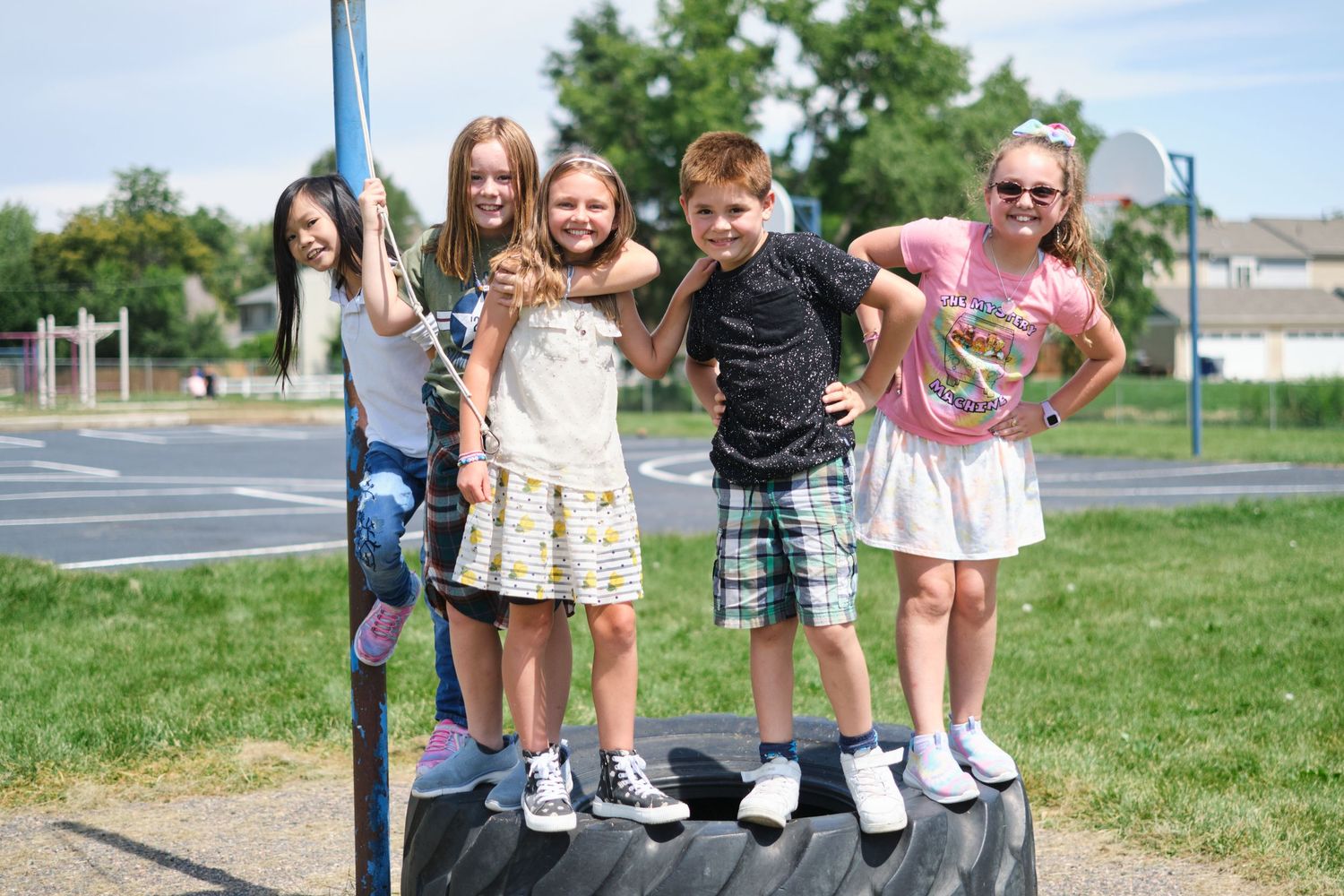 A group of children stand together smiling on a raised platform outdoors near a grassy area on a sunny day.