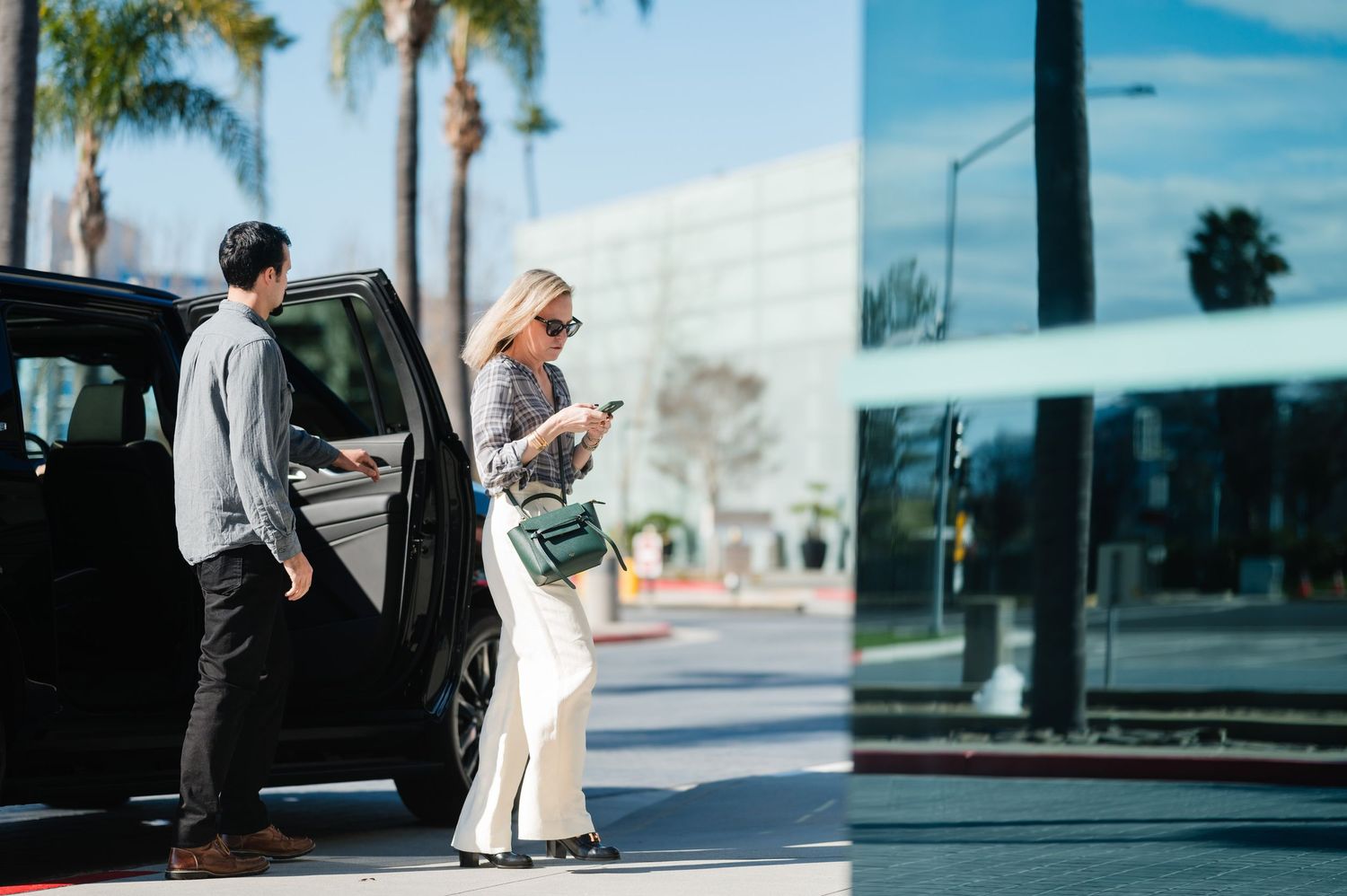 A person in gray stands by a black car while another checks their phone on a sunny street with palm trees.