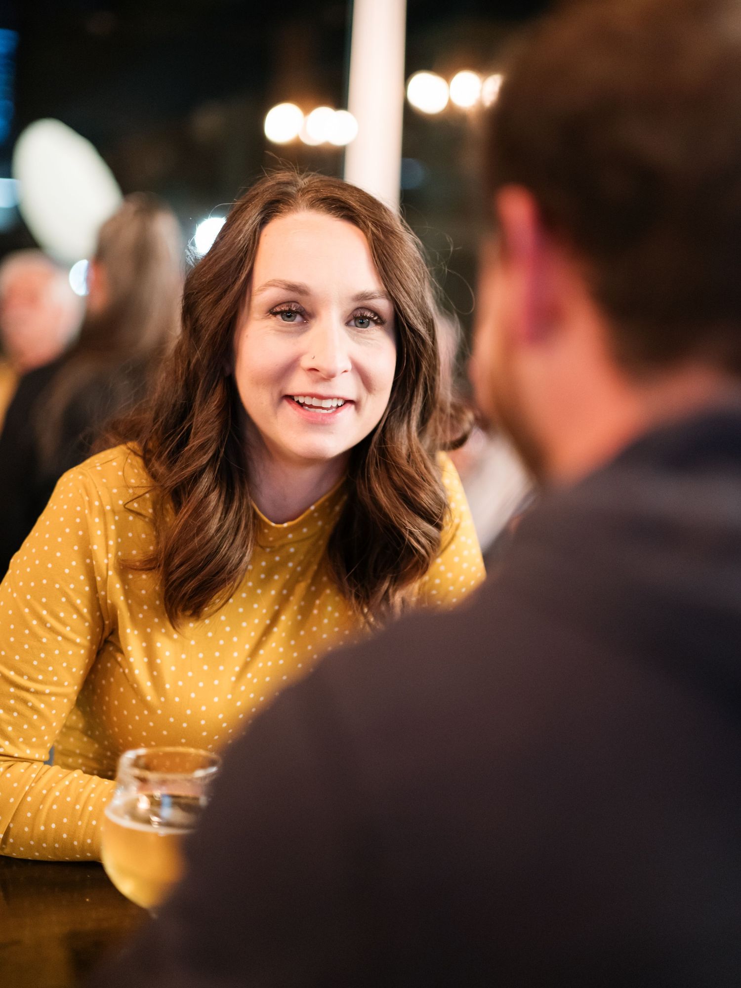 A person in a yellow sweater smiles warmly while engaged in conversation at a restaurant or bar.