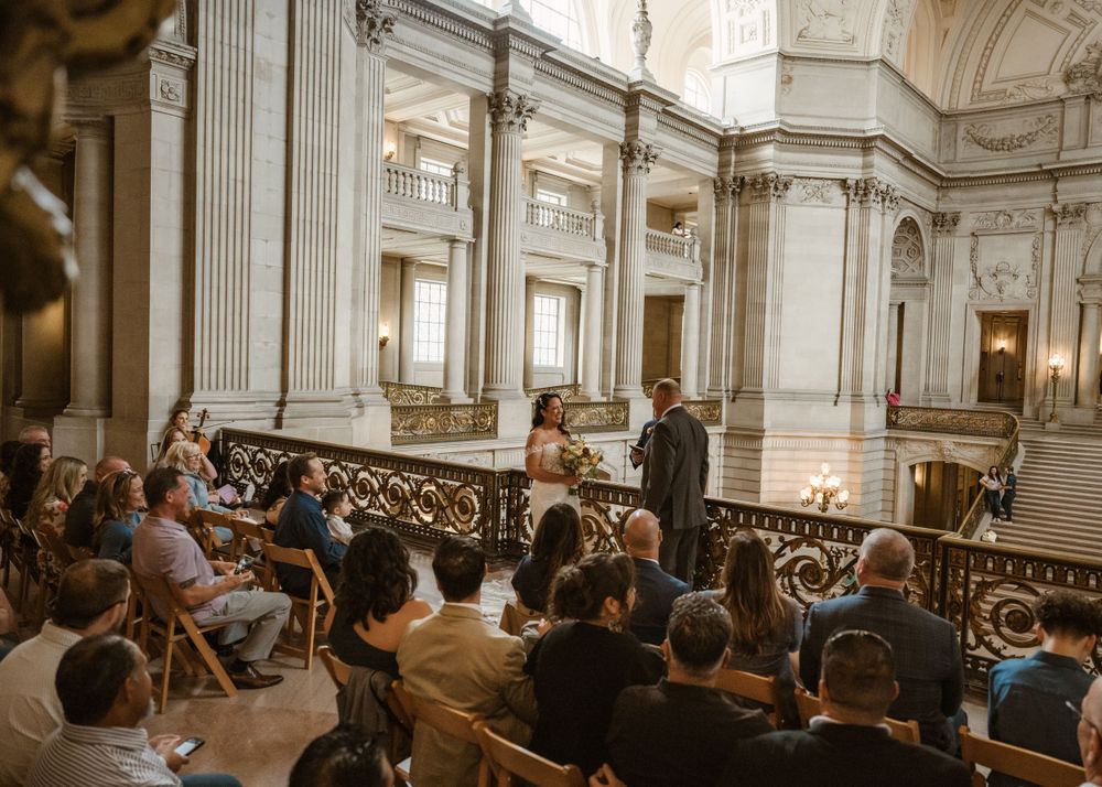Wedding ceremony in progress inside ornate marble hall with crowd seated.