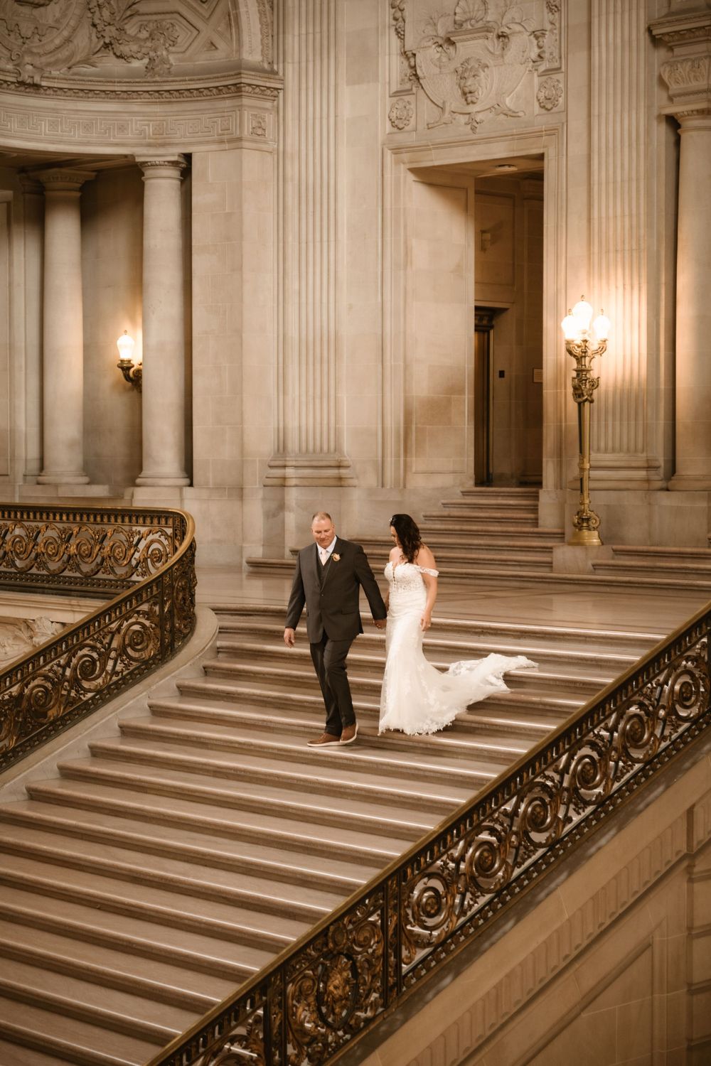 A couple descends a grand marble staircase with ornate gold railings in a historic building.