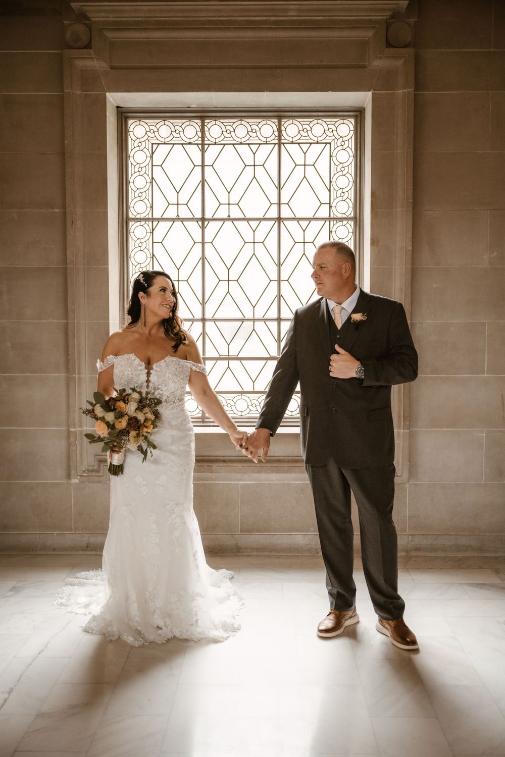 A couple poses by a geometric window in an elegant building, with the bride holding a floral bouquet.
