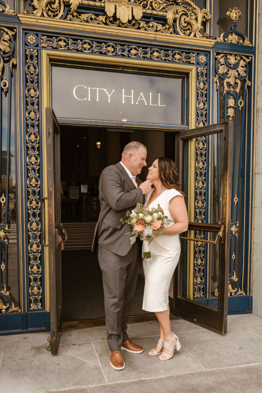 A couple shares a tender moment outside City Hall's ornate entrance doors.