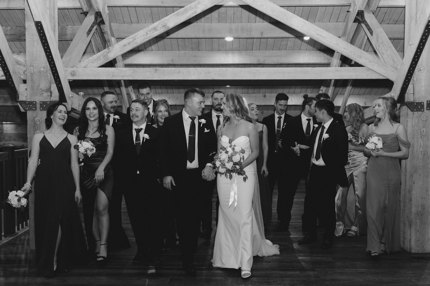 Black and white photo of a wedding party walking together inside a rustic wooden barn venue.