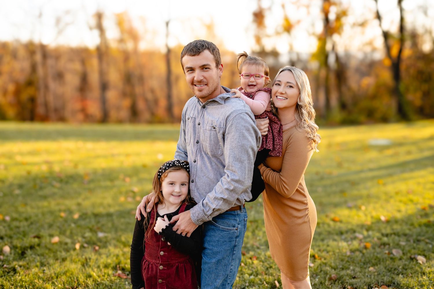 A happy family of four smiles together in a fall field during golden hour sunset.