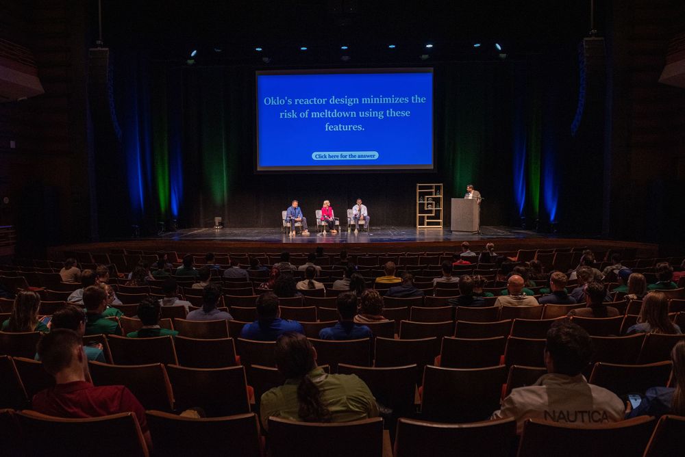 Theater audience watching a panel discussion on stage with projected text display.