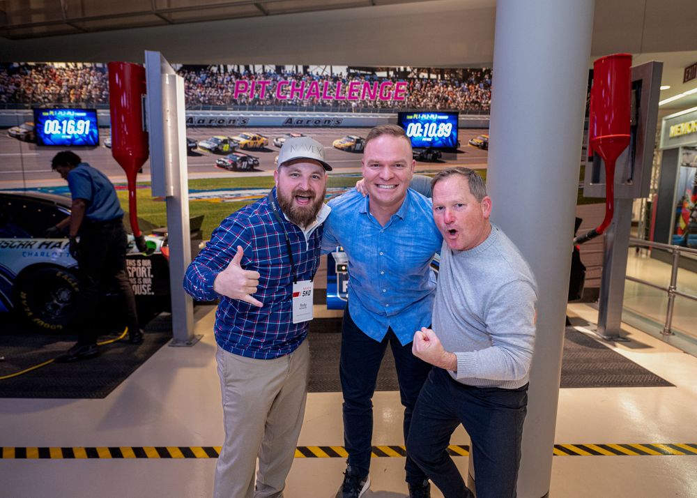 Three people pose together in front of racing simulator display at event.