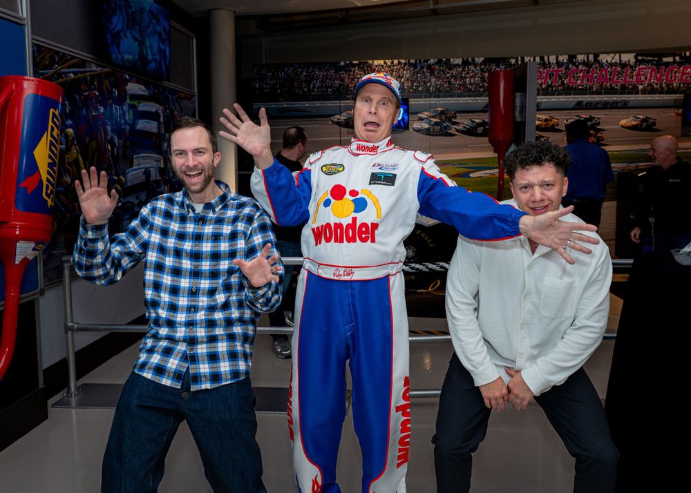 Three enthusiastic people pose with a colorful racing uniform display at a motorsports event.