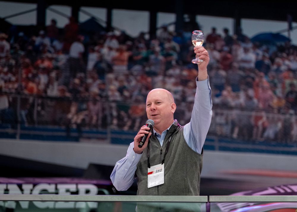 Speaker addresses crowd while holding microphone and raising water bottle at outdoor stadium event.