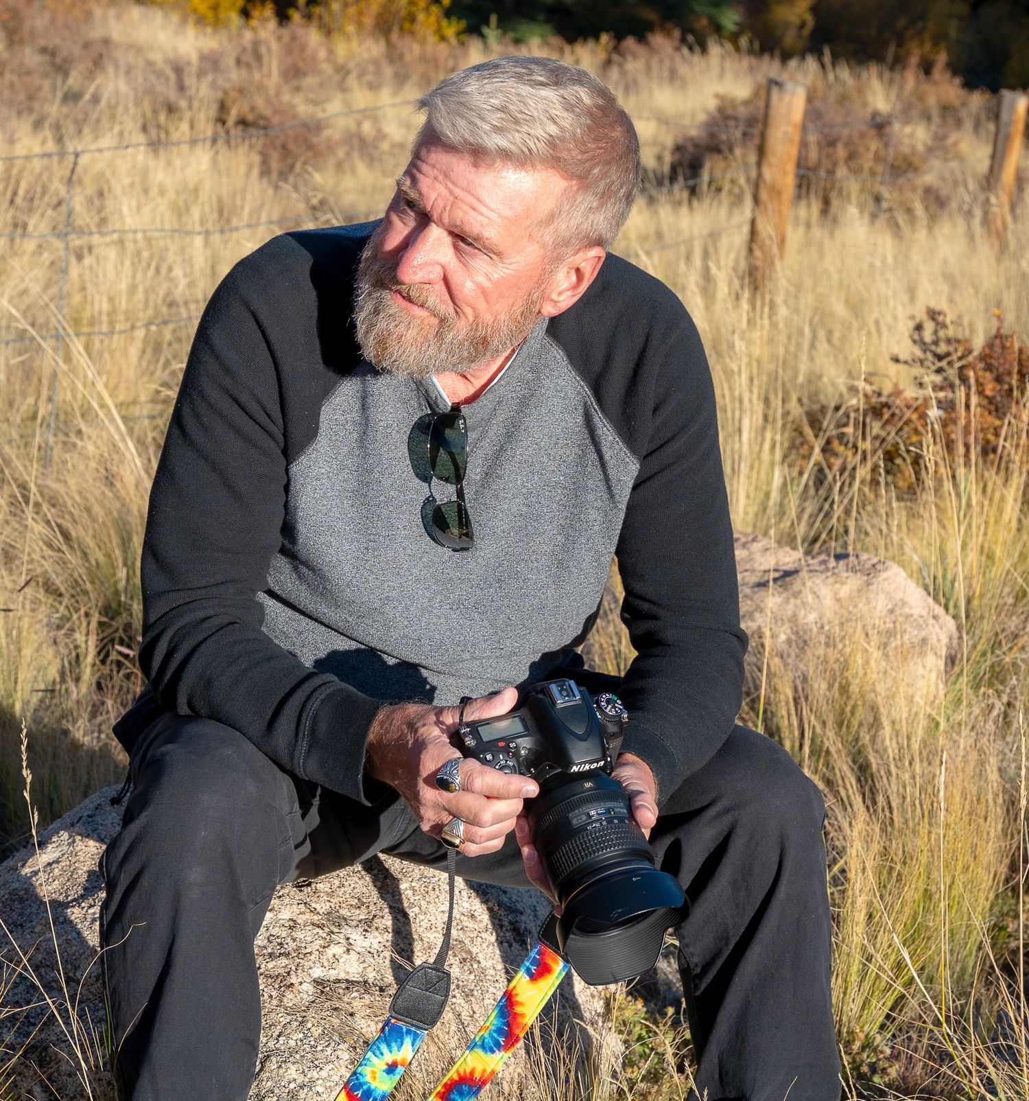 Photographer with beard sitting on rock holding camera in grassy field.