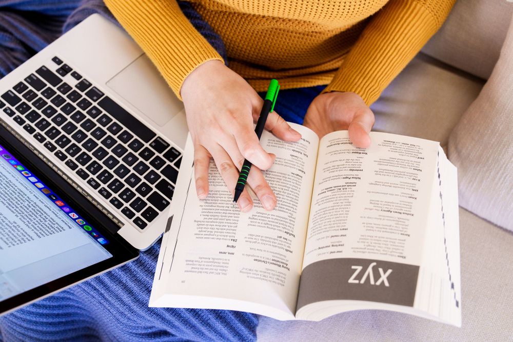 Hands holding pen and book next to laptop while studying.