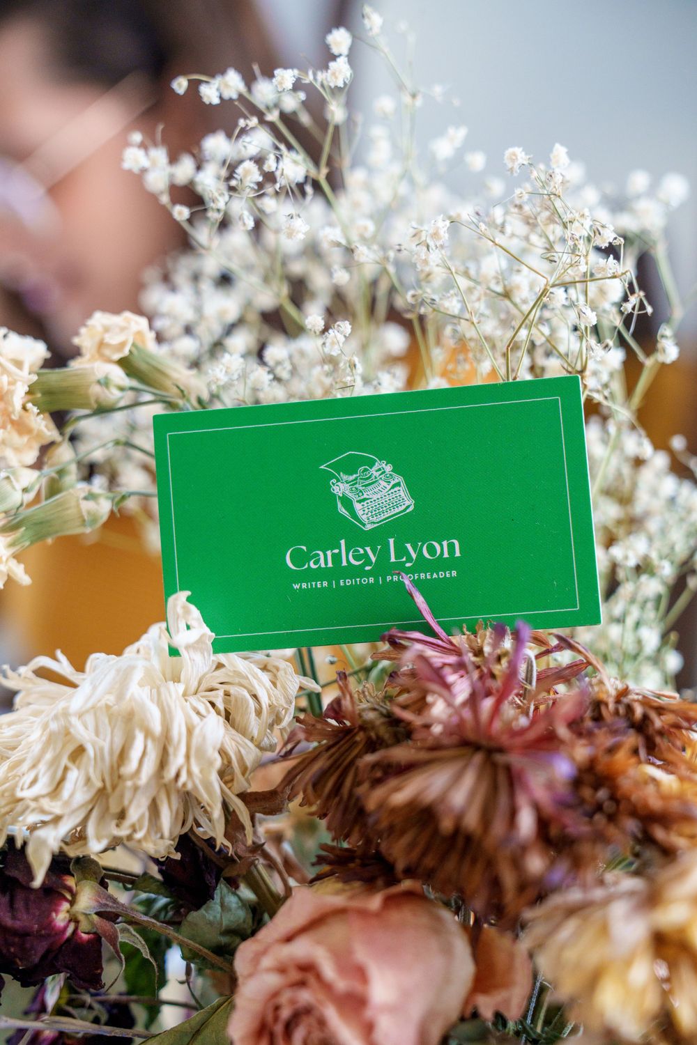 Green Carley Lyon business card displayed among delicate white baby's breath flowers and dried roses.