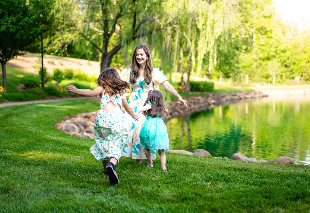 Family photograph near me captured a tender moment of a mother playing with her children in a park with a pond on the background.