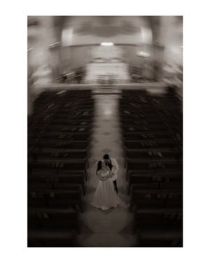 Artistic black and white photo of newlyweds standing in empty church aisle surrounded by wooden pews.