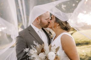 A romantic wedding moment as a couple shares a kiss under a flowing white veil in natural sunlight.