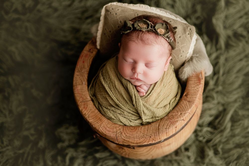 Sleeping newborn wrapped in olive green swaddle resting in wooden bowl with floral headpiece.