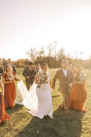 Wedding party walking together across a sunlit field with bridesmaids in rust-colored dresses.