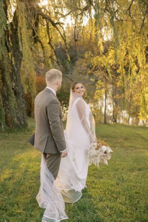 Newlyweds pose under weeping willow trees during golden hour with flowing veil and bouquet.