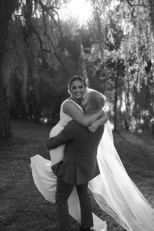 A joyful wedding embrace captured in black and white under weeping willow trees.