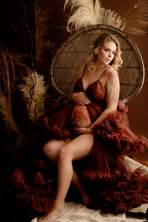 Dramatic maternity portrait in burgundy dress seated on peacock chair with feather accents.